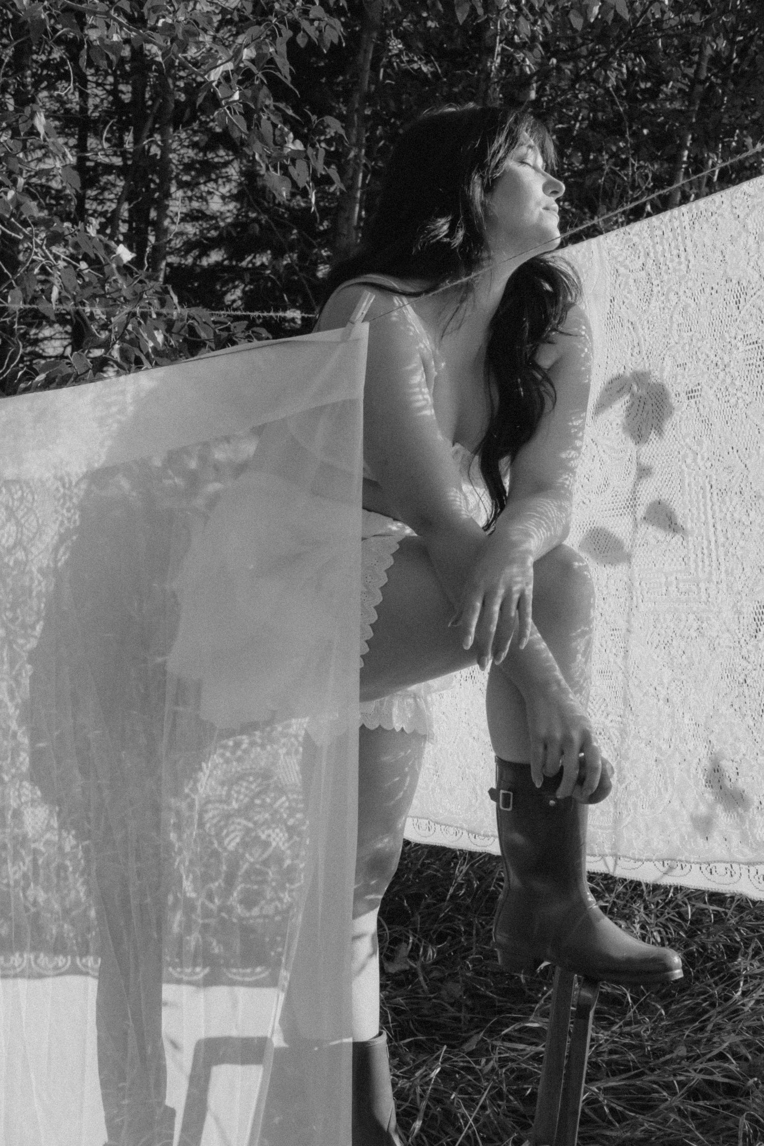 Black and white photo of a woman in lace top and boots leaning against hanging white fabric outdoors.