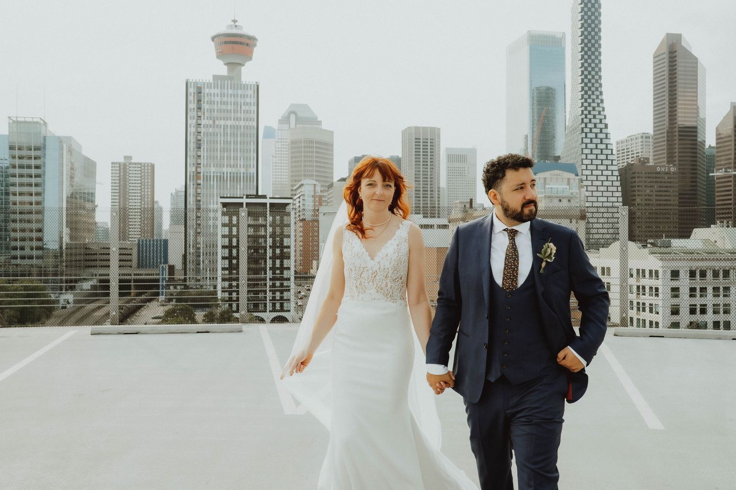 Couple walks hand-in-hand on rooftop with Vancouver skyline backdrop, bride in white gown, groom in navy suit.