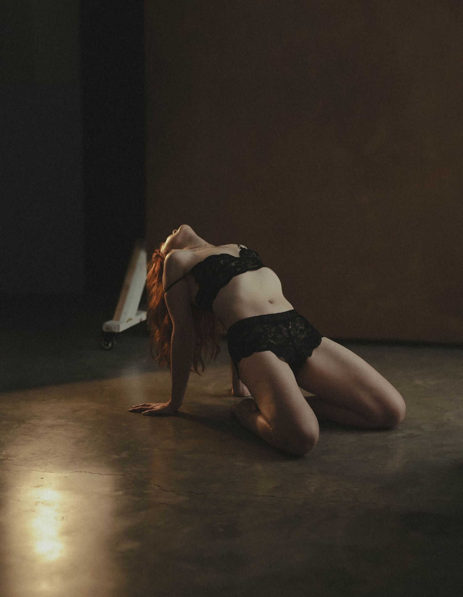 Red-haired woman in black lingerie posing gracefully on a concrete floor in moody dramatic lighting.