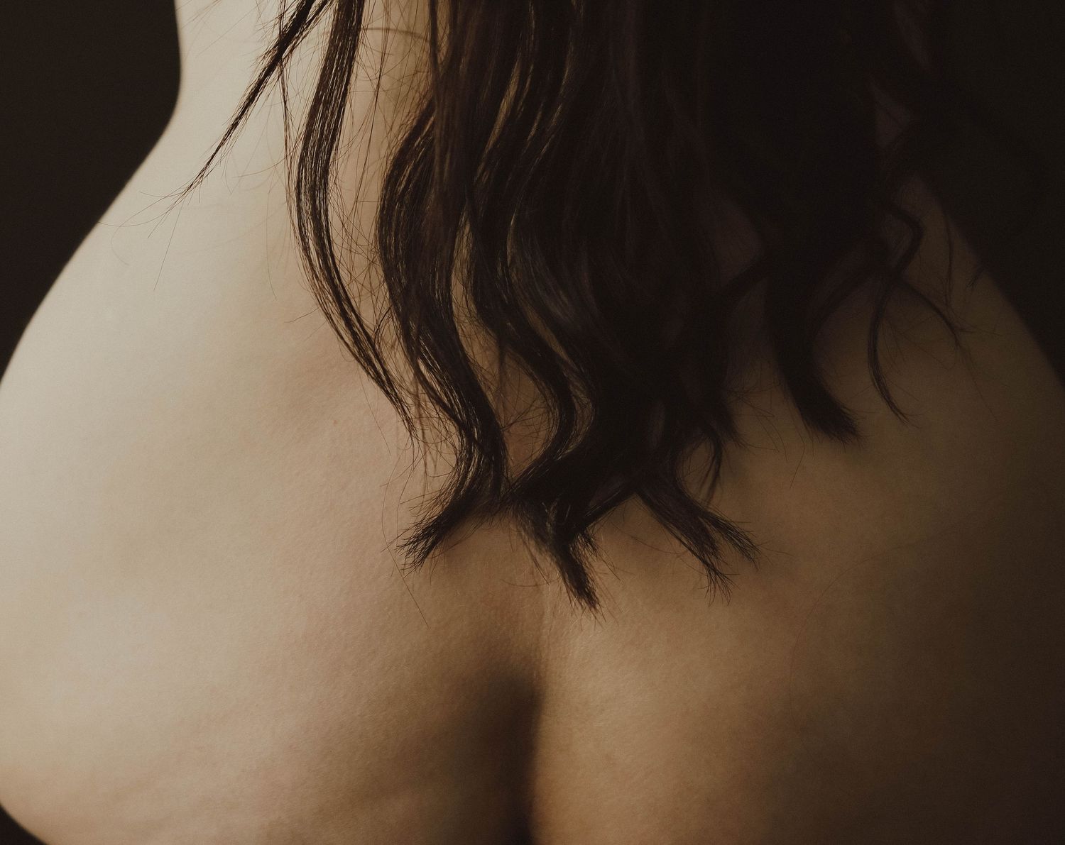 Close-up of a person's bare upper back and shoulders with dark wavy hair cascading down.