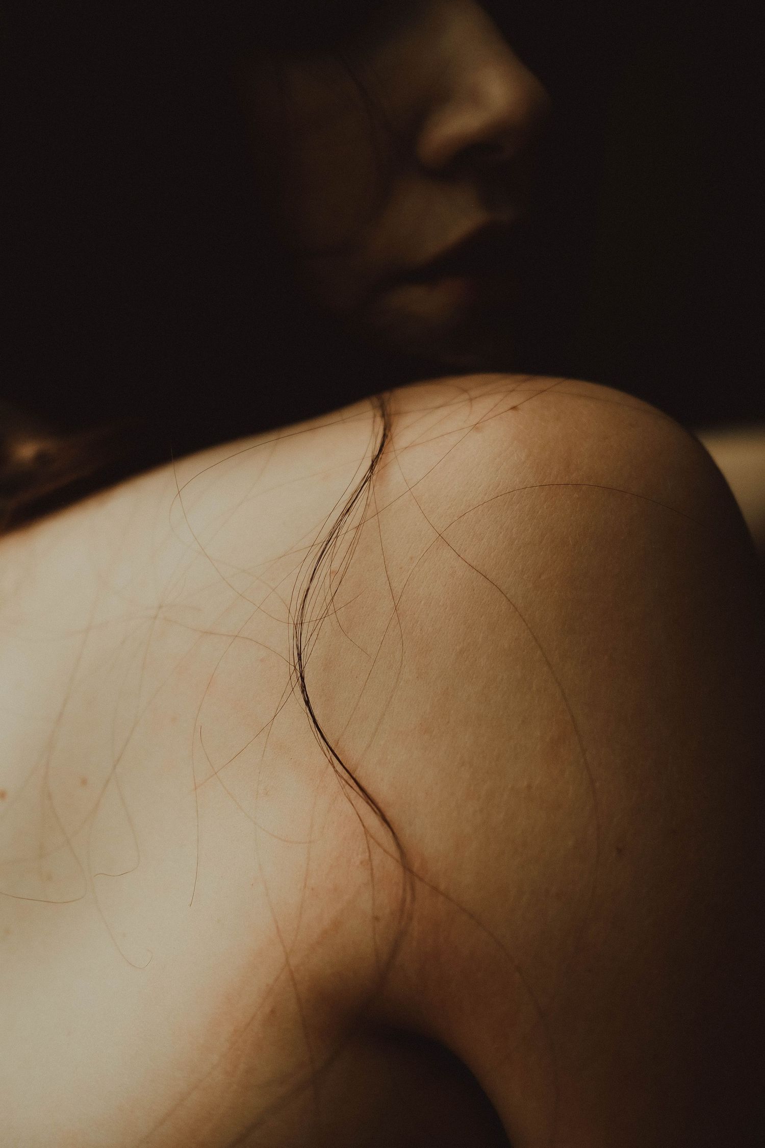 Close-up of a bare shoulder with strands of hair draped across skin in dramatic moody lighting.
