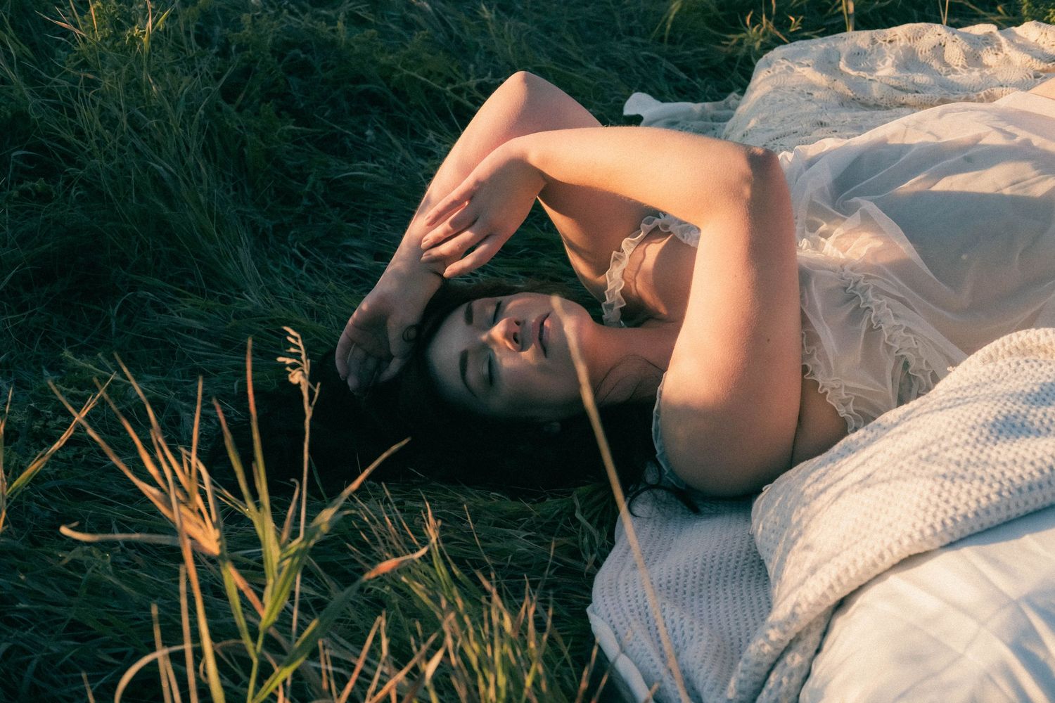Woman in white dress lying in green grass field, shielding eyes from golden sunlight in serene outdoor setting.