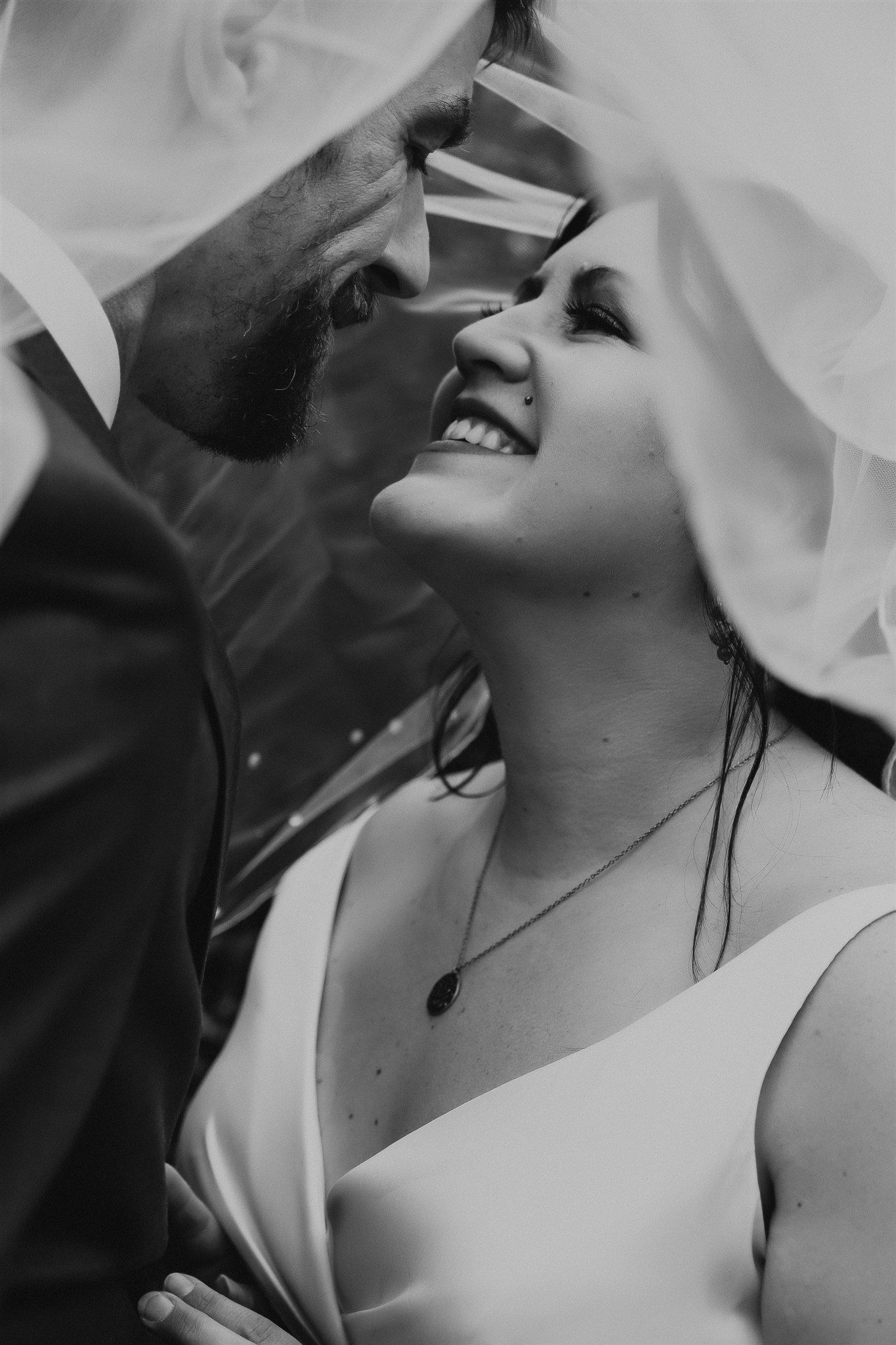Black and white wedding portrait of a smiling bride under a veil with groom leaning in close.