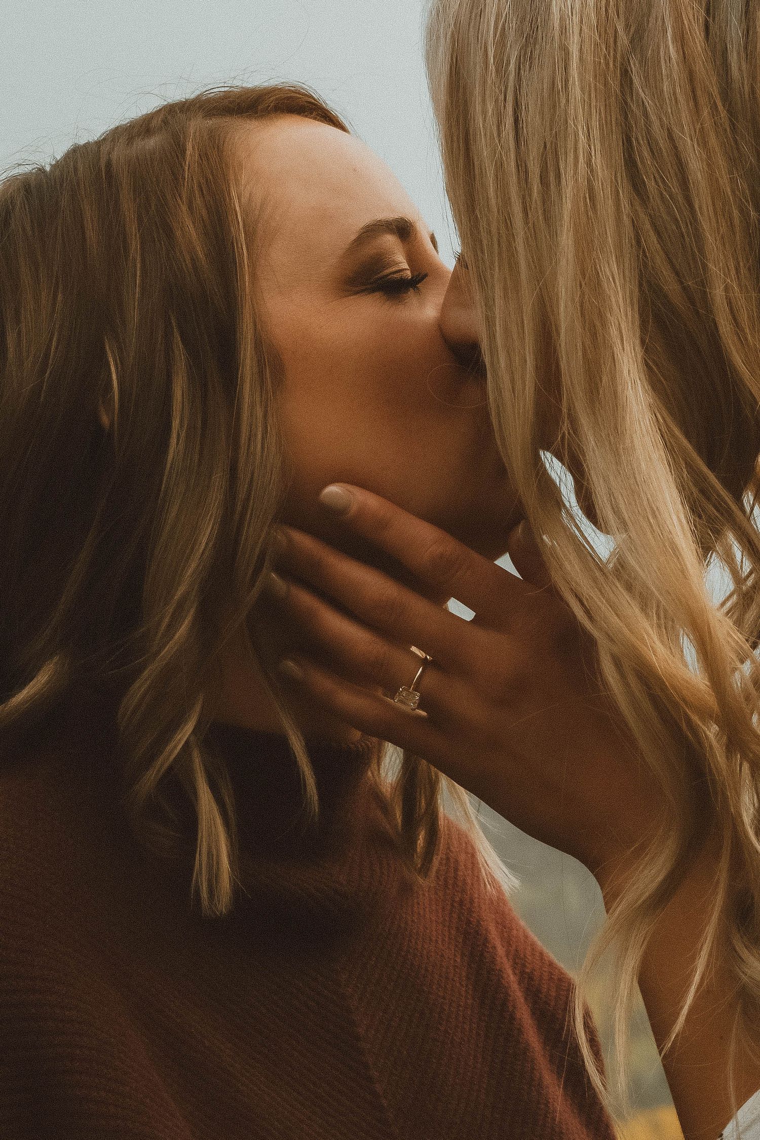 Two blonde women share a tender kiss outdoors, one wearing a delicate gold ring.