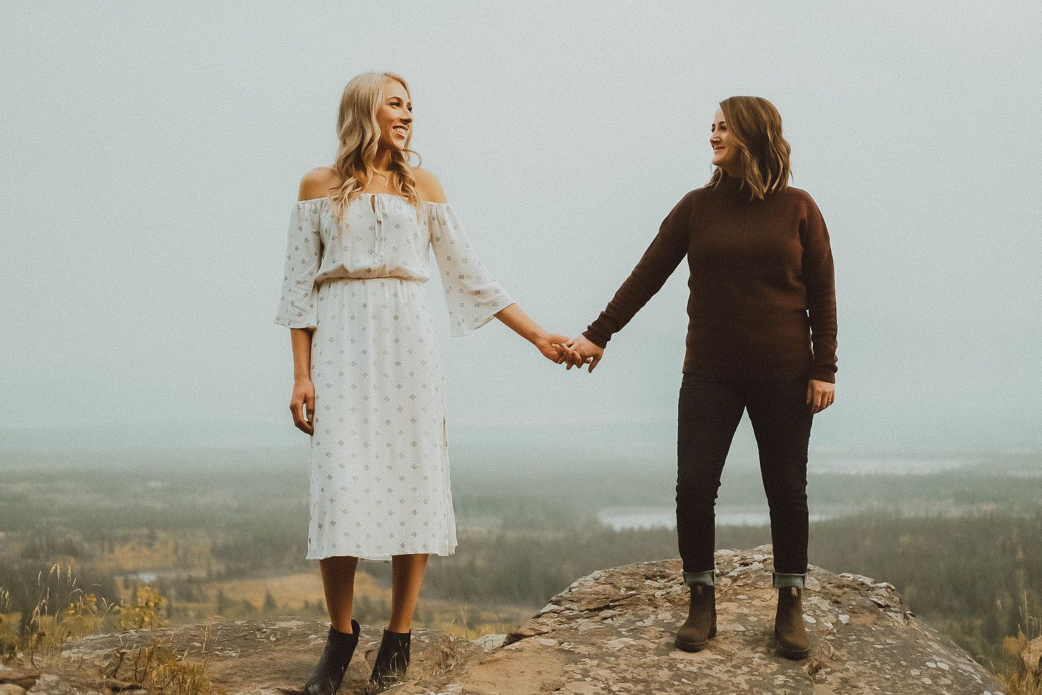 Two women hold hands on a rocky mountain overlook on a misty overcast day.