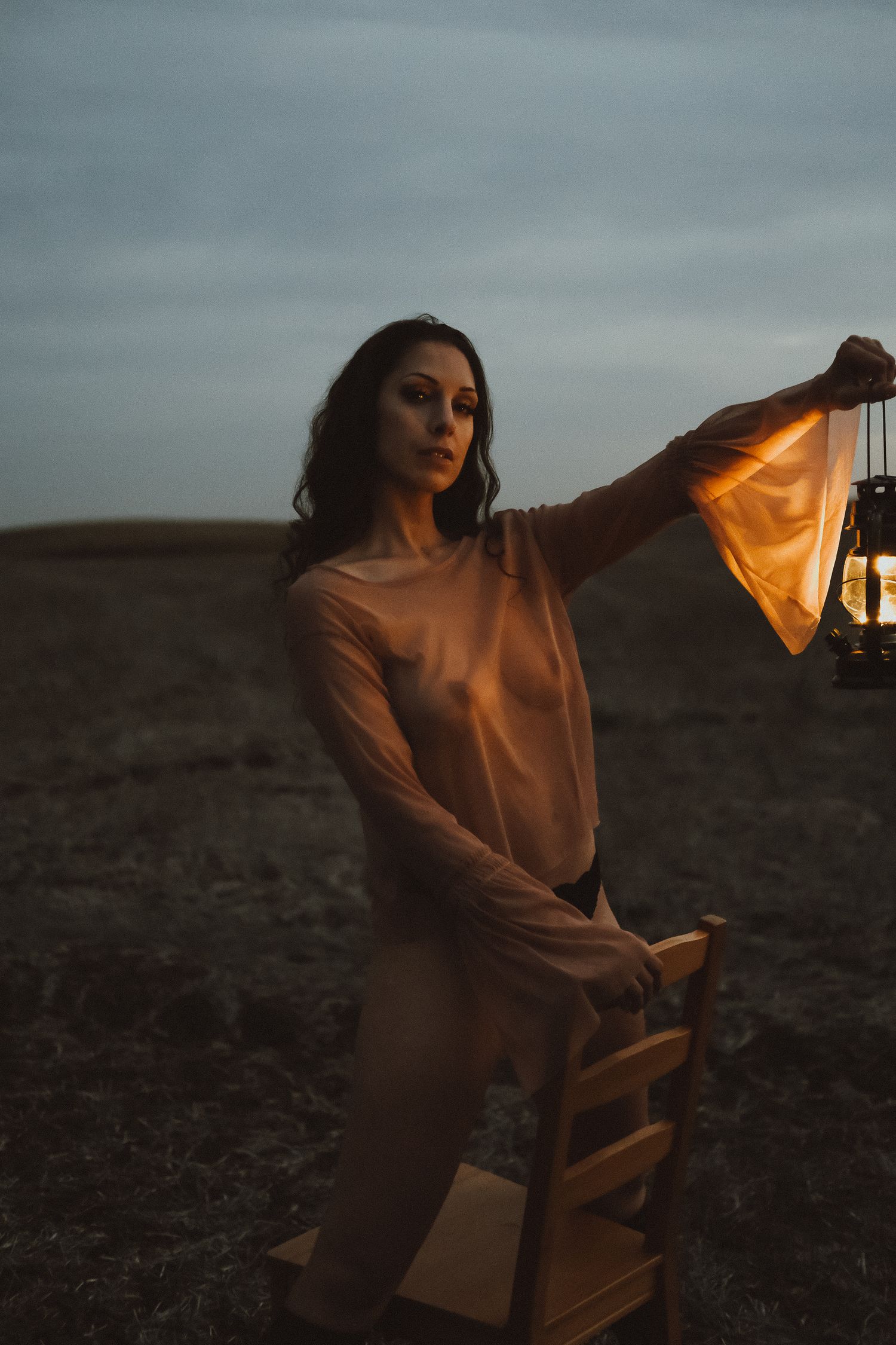 Woman in sheer top holding vintage lantern outdoors at dusk near wooden chair in open field.