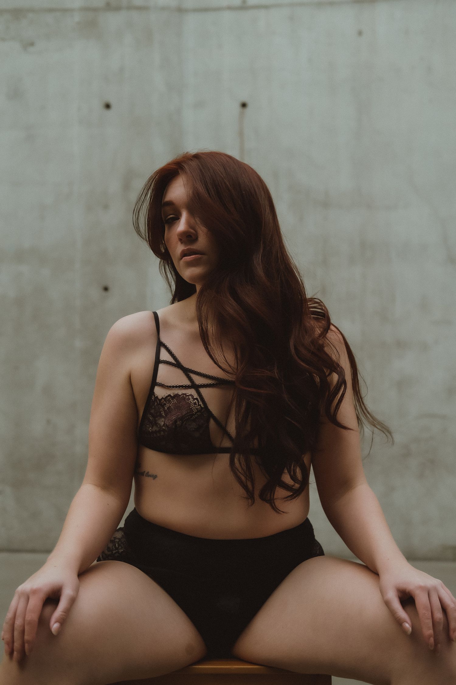 Young woman with long auburn hair posing in black lace lingerie against concrete wall.