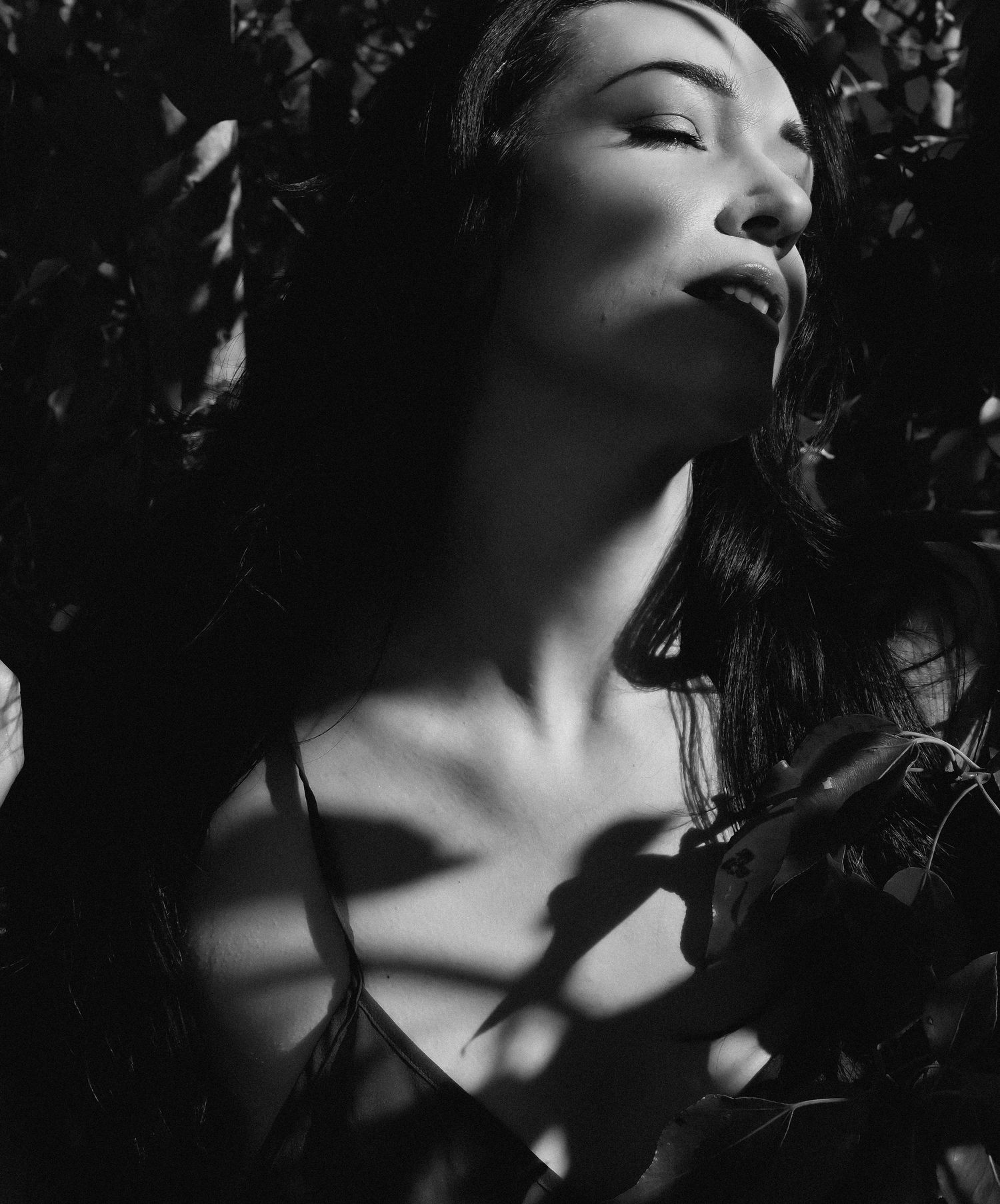 Black and white portrait of a woman with closed eyes among dark leaves, dramatic shadows highlighting her neck and face.