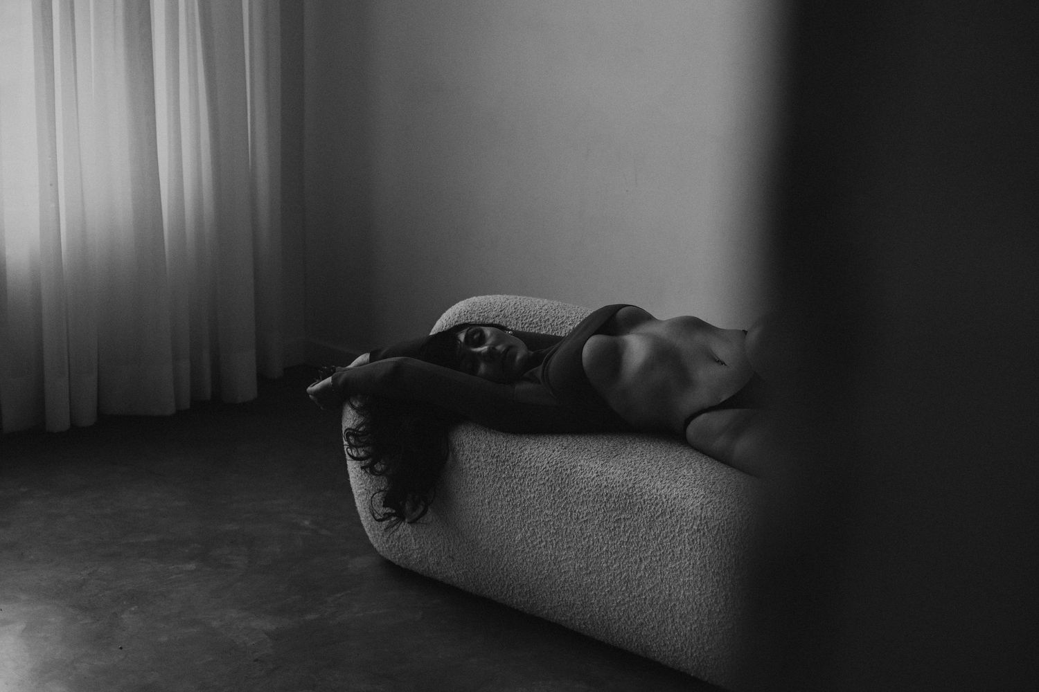 Black and white artistic photo of woman lying on white boucle sofa near sheer curtains.