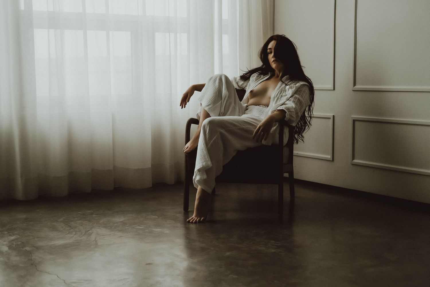 Woman in white outfit relaxing in armchair by sheer curtained window on concrete floor.