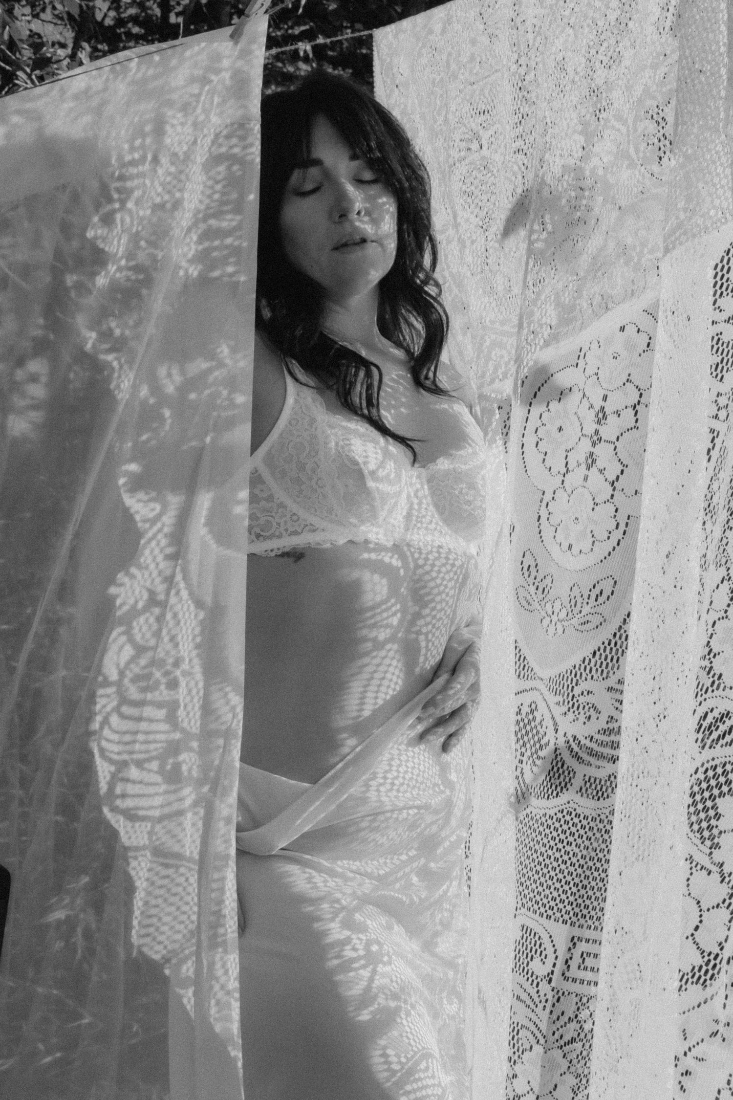 Black and white photo of woman in lingerie behind lace curtains outdoors.