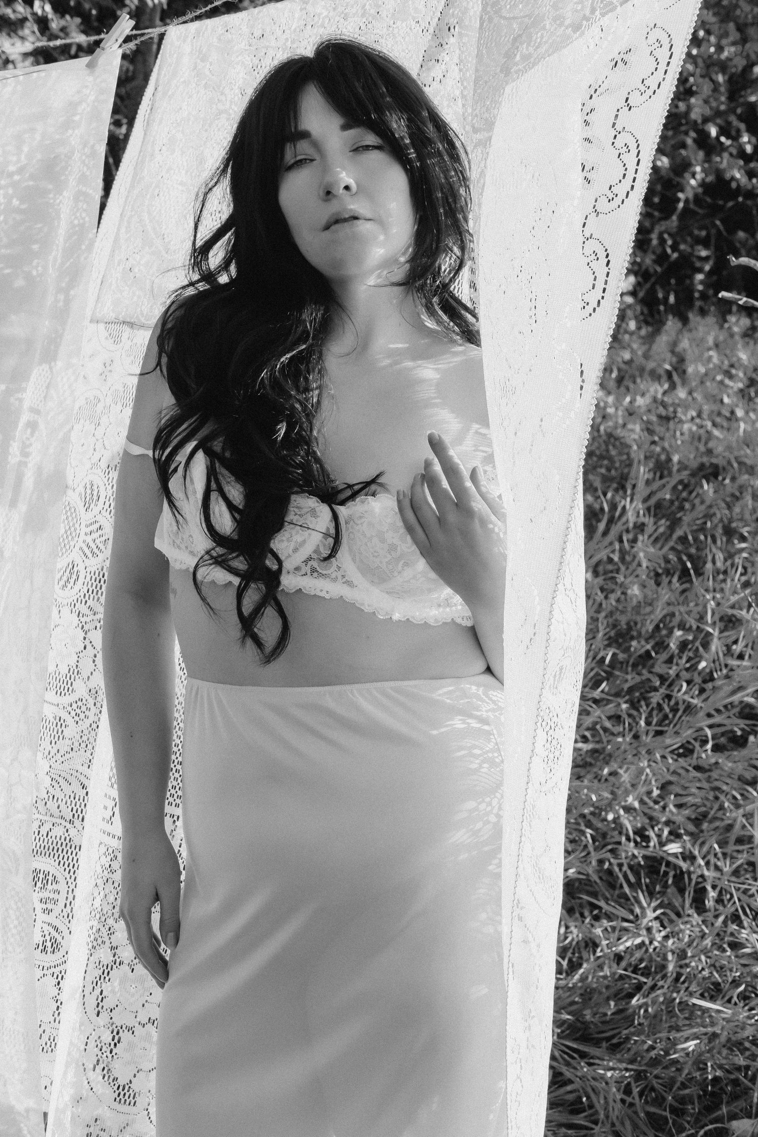 Black and white portrait of a woman in lace bralette and satin skirt standing between hanging white linens.