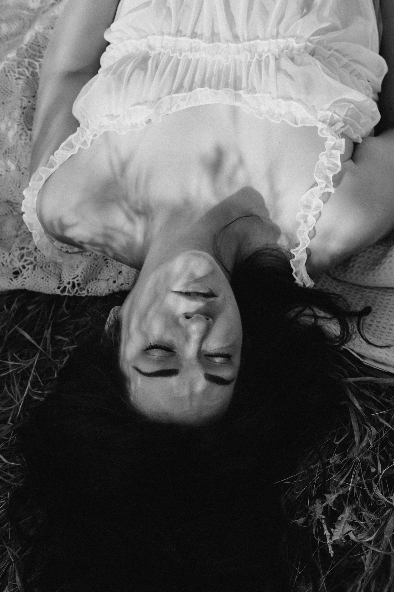 Black and white photo of a woman in white dress lying on grass with eyes closed, shadows across her face.