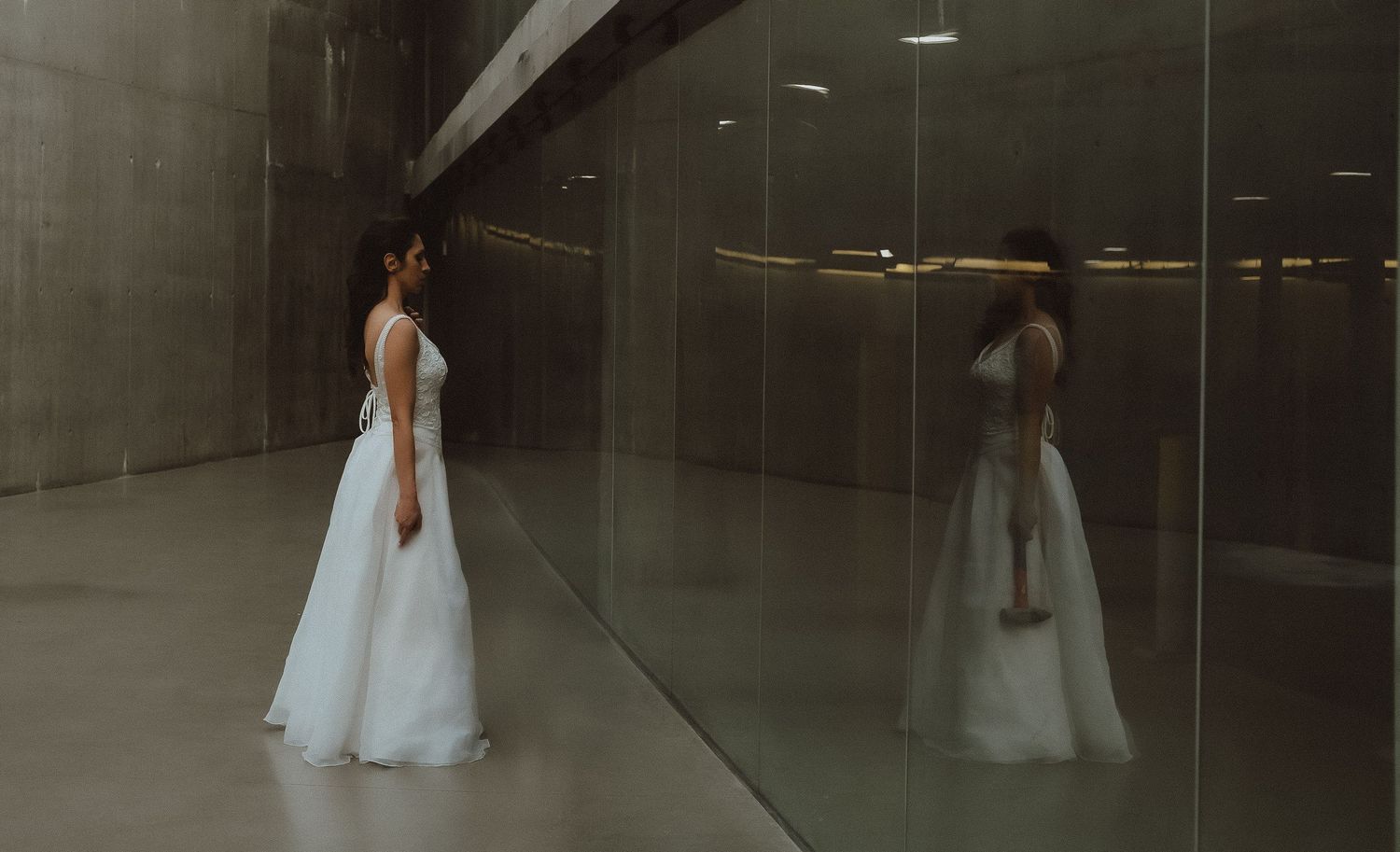 Bride in white gown stands before mirrored wall reflecting her elegant silhouette in a modern concrete space.