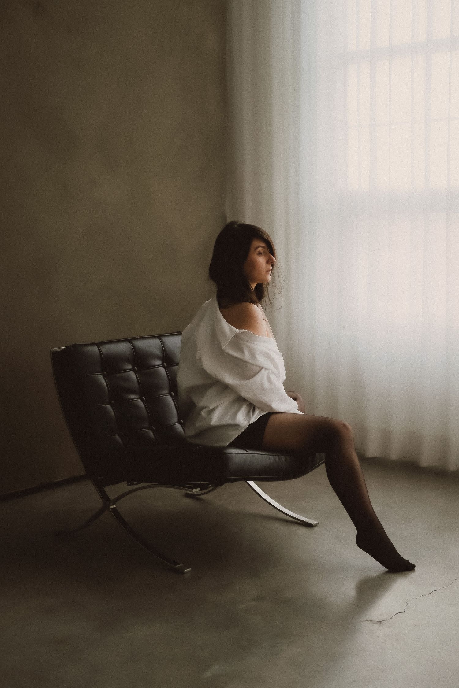 Woman in white off-shoulder top seated on leather Barcelona chair by sheer curtained window.