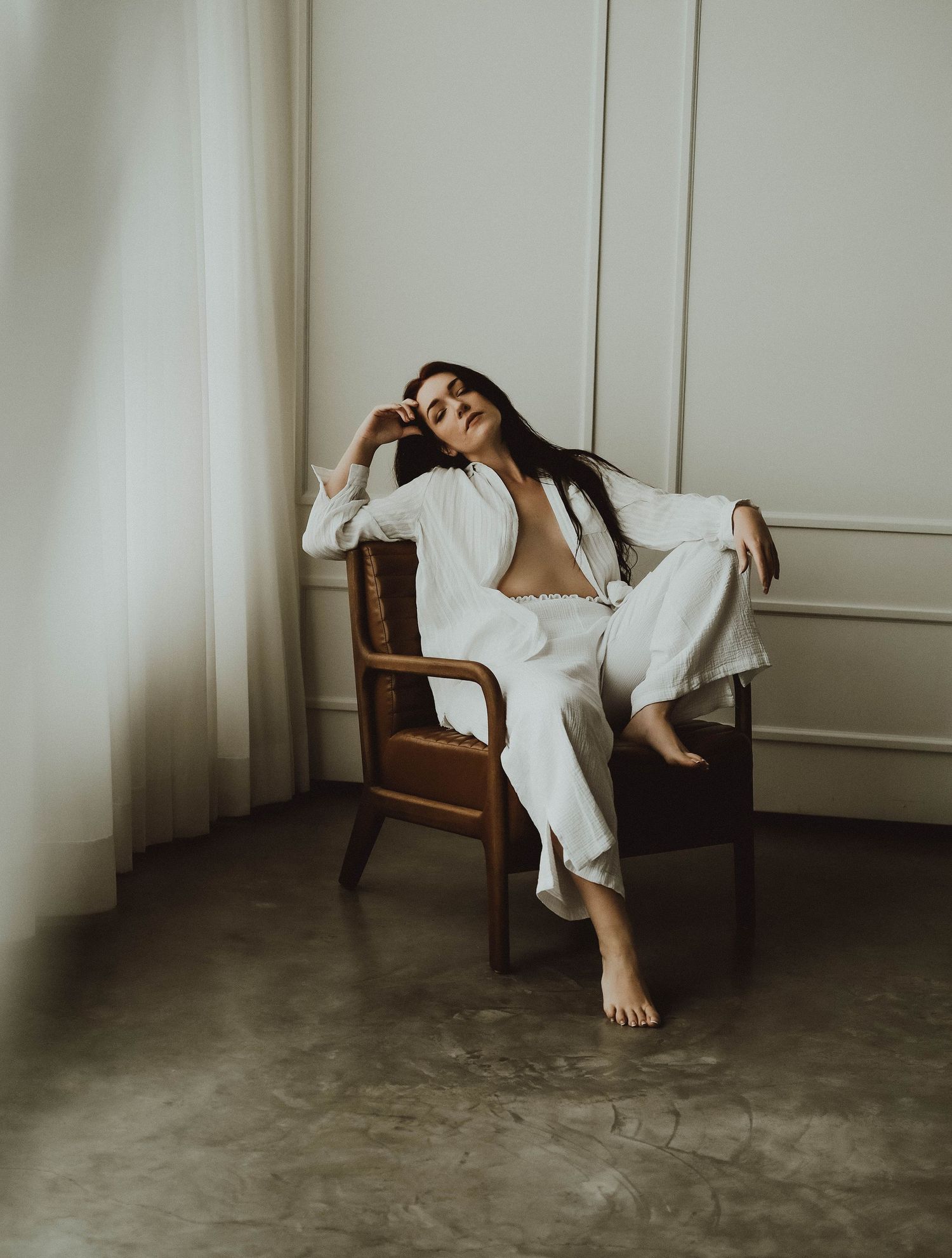 Woman in white linen suit relaxing barefoot on a wooden armchair in a minimalist room.