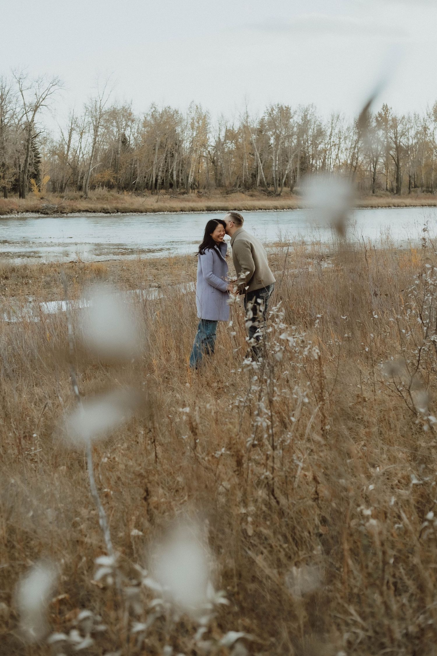 Couple embracing in golden autumn field near river, surrounded by dry grass and bare trees on overcast day.