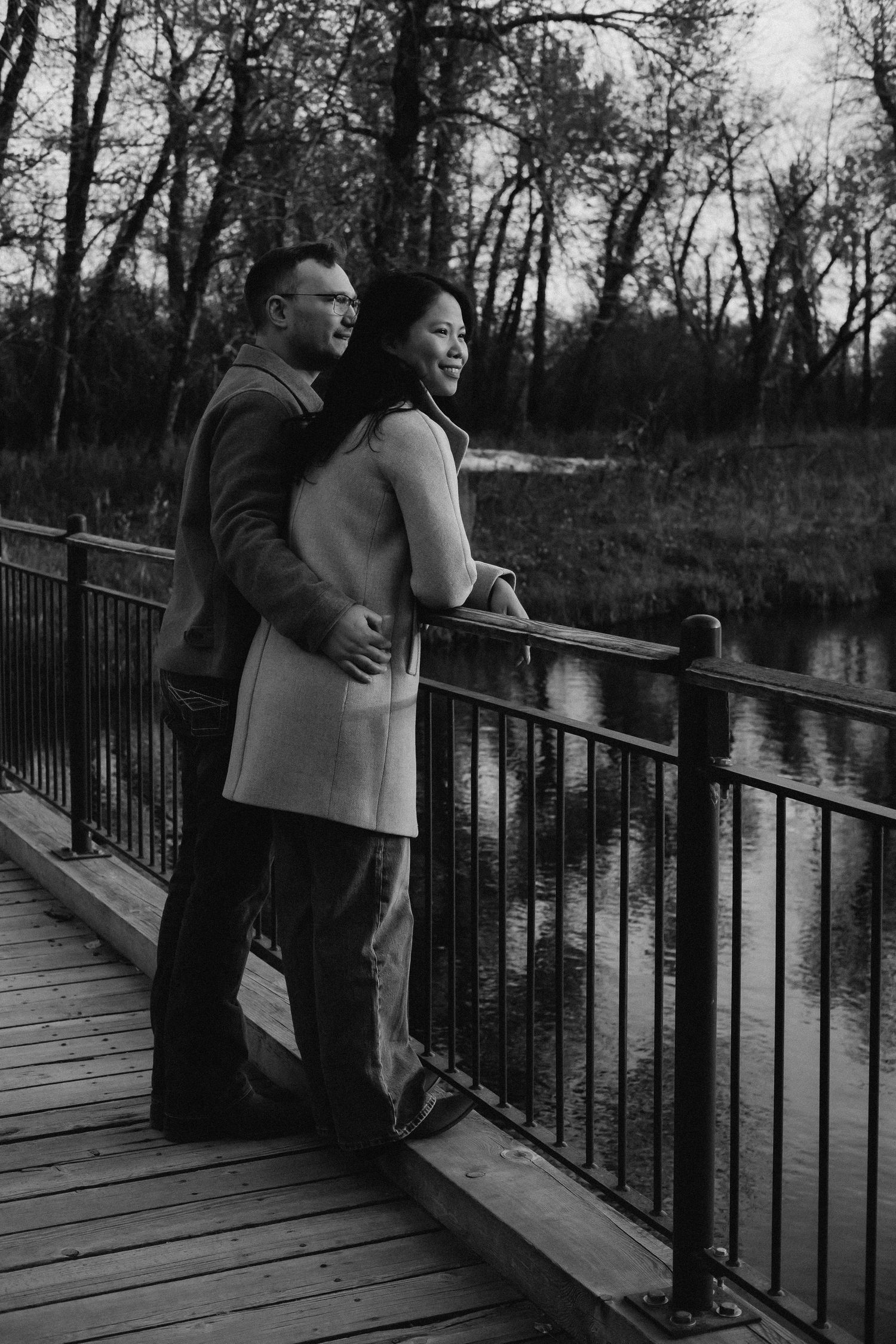 Black and white photo of a couple embracing on a wooden bridge overlooking a serene river surrounded by trees.