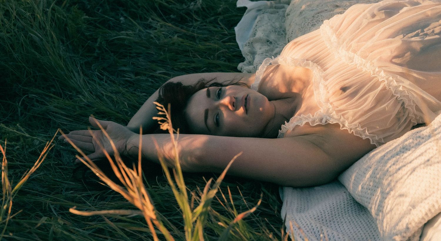 A young woman with blonde hair lies peacefully in green grass, wearing a white lace dress in soft natural light.