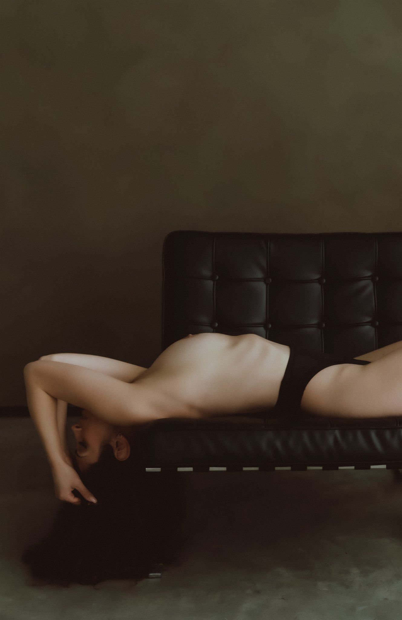 Artistic nude figure lying face-down on black leather sofa in dramatic moody lighting.