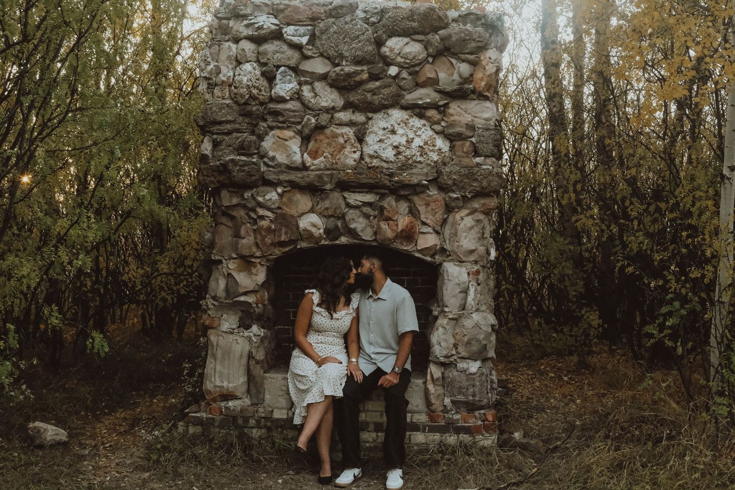 Couple poses romantically in front of a rustic stone fireplace surrounded by autumn trees in an outdoor setting.