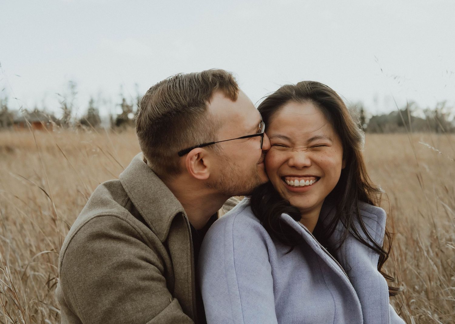 A man kisses a laughing woman on the cheek while sitting in a golden field outdoors.