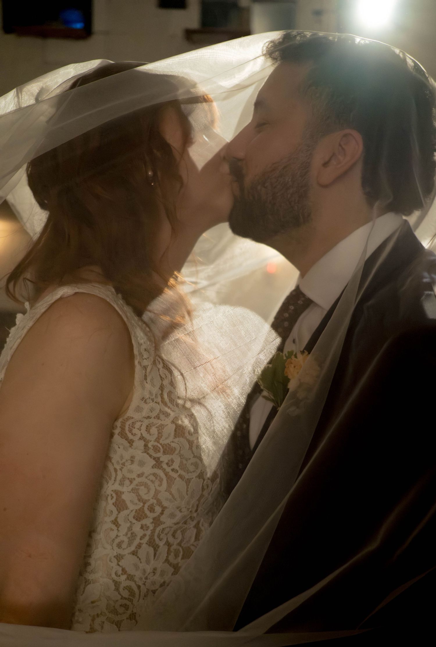 Bride and groom sharing a romantic kiss under a wedding veil, soft warm lighting highlighting their special moment.