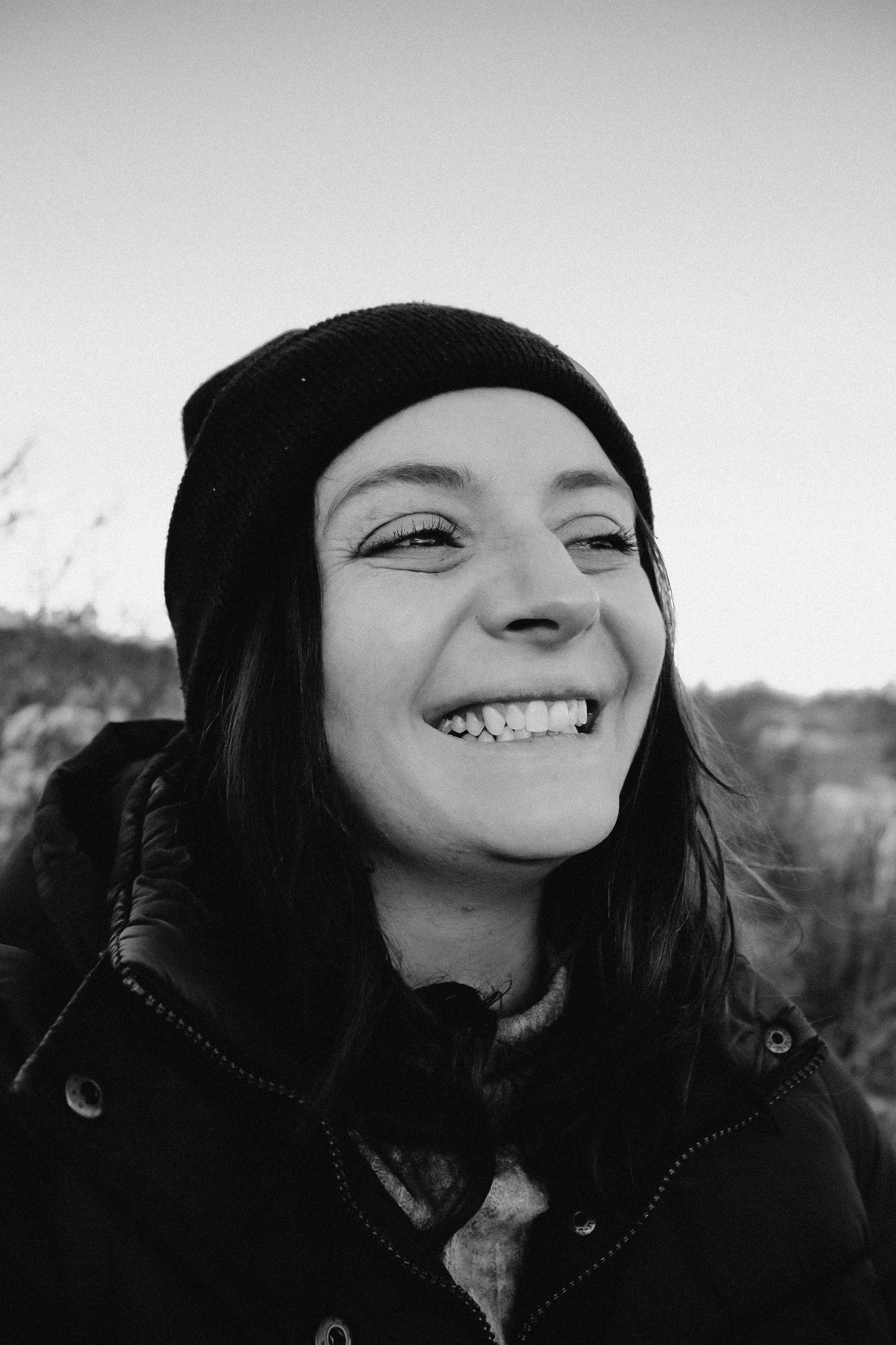 Black and white portrait of a smiling young woman wearing a beanie hat and winter jacket outdoors.