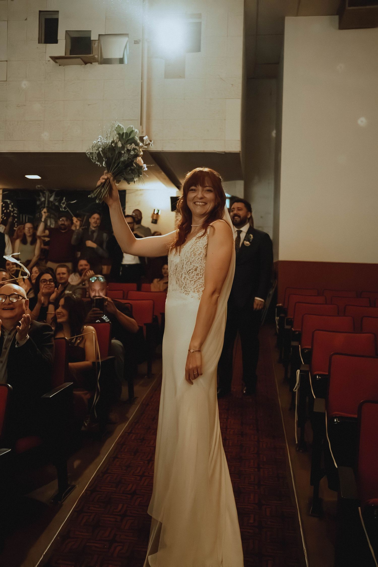 Bride in white lace gown raises bouquet while walking down aisle in venue with red seats.