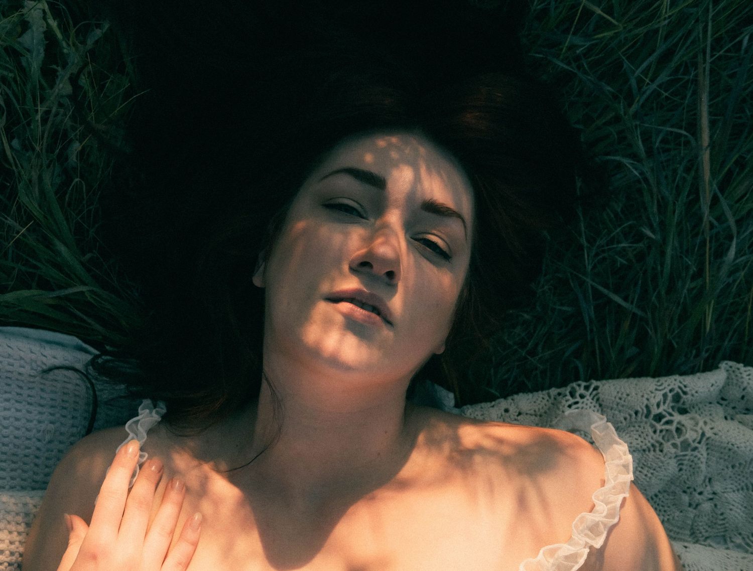 Close-up of a dark-haired woman lying in grass with dappled sunlight across her face and white ruffled top.