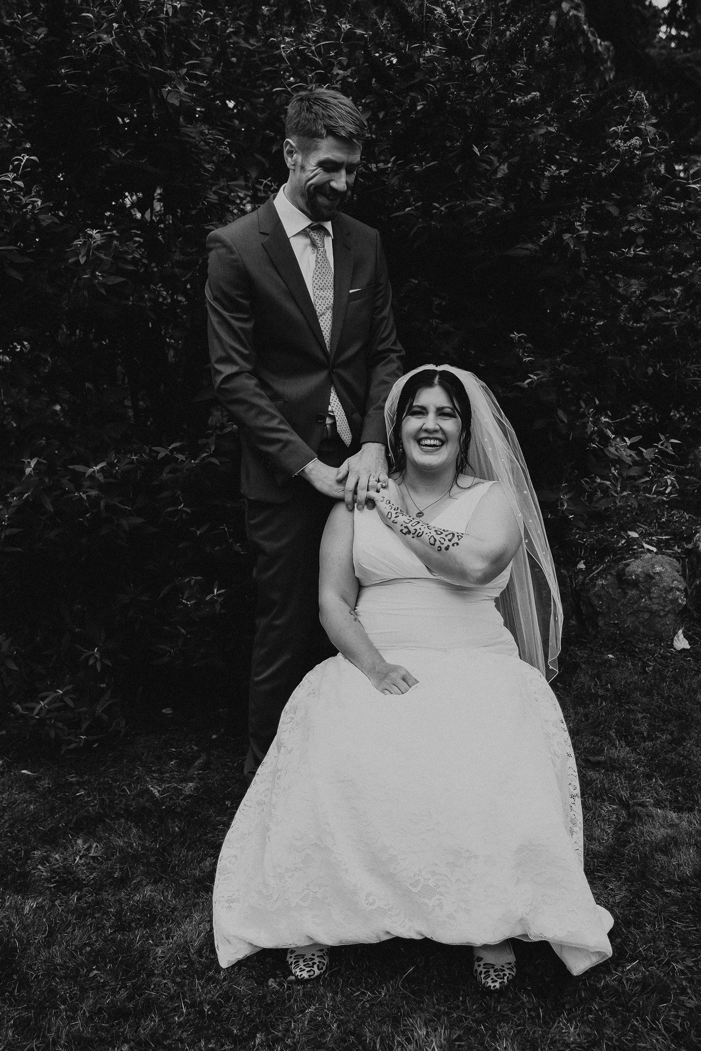 Black and white portrait of smiling bride seated in wedding gown with veil, groom standing behind her outdoors.