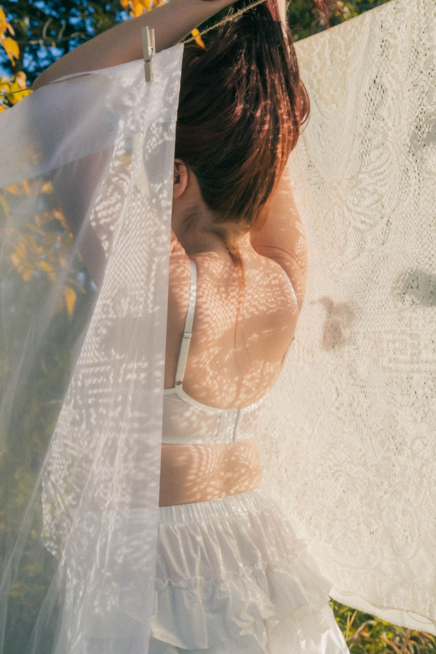 Woman in white lingerie partially hidden behind lace curtains on clothesline in autumn sunlight.