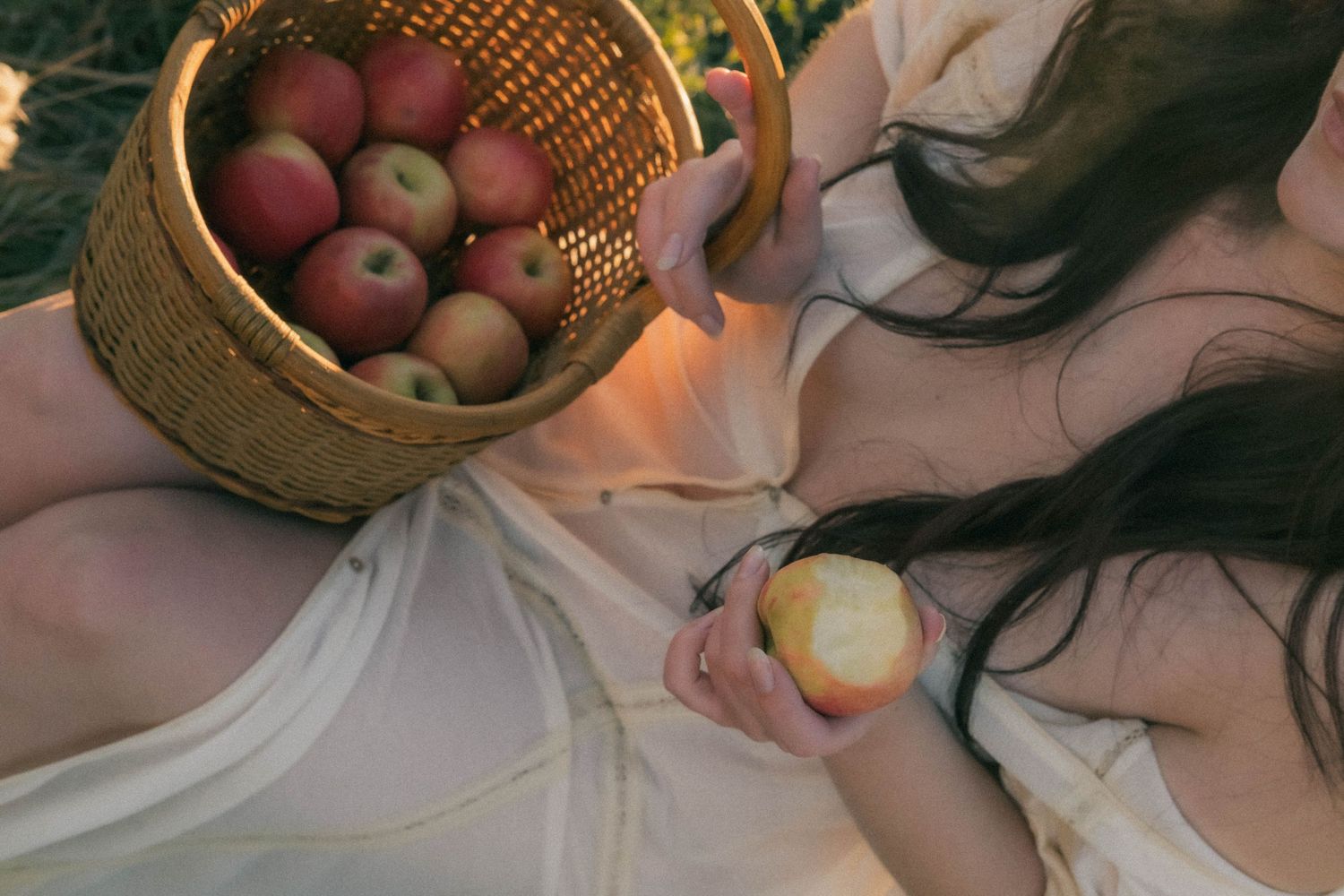 Woman in white dress lying outdoors, holding a bitten apple beside a wicker basket filled with fresh red apples.