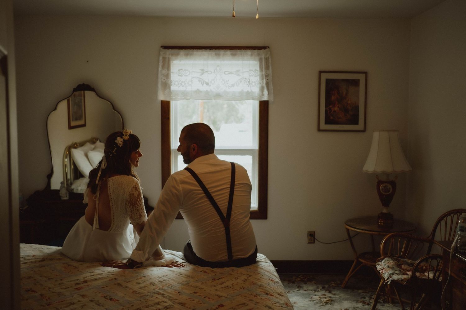 Bride and groom sitting on bed gazing out window, romantic wedding day moment in vintage bedroom.