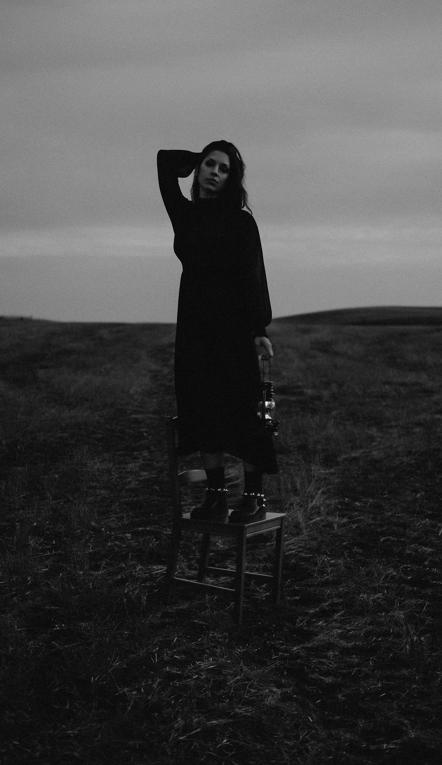 Black and white photo of a woman in dark clothing standing on a stool in an open field under a cloudy sky.
