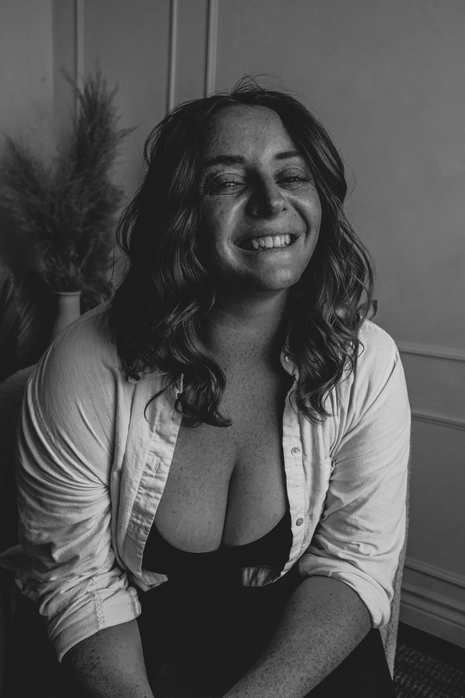 Black and white portrait of a smiling woman with wavy hair wearing an open white shirt, sitting indoors.