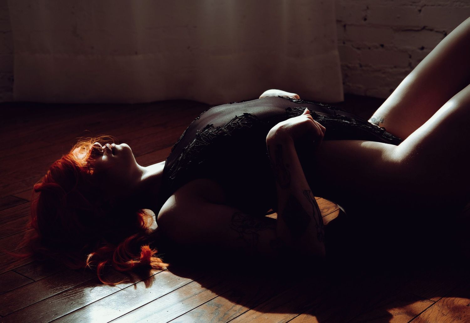 A redheaded woman in black lingerie poses dramatically on a hardwood floor in moody, atmospheric lighting.