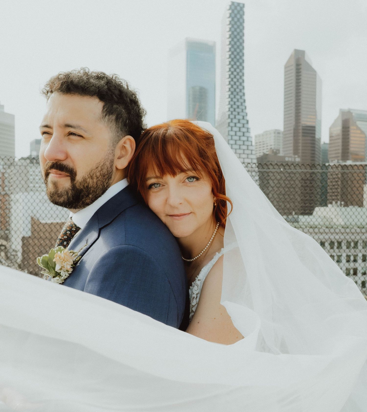 Bride and groom pose romantically on a rooftop with city skyline backdrop, bride in veil leaning on groom's shoulder.
