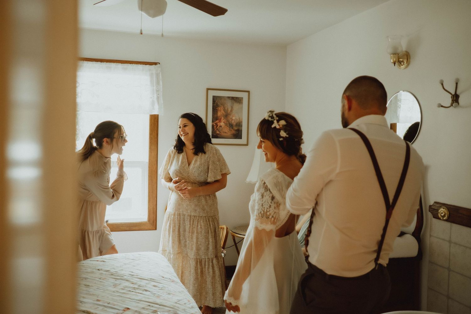 Bridesmaids help bride get ready in cozy bedroom before wedding ceremony, wearing elegant white and cream dresses.