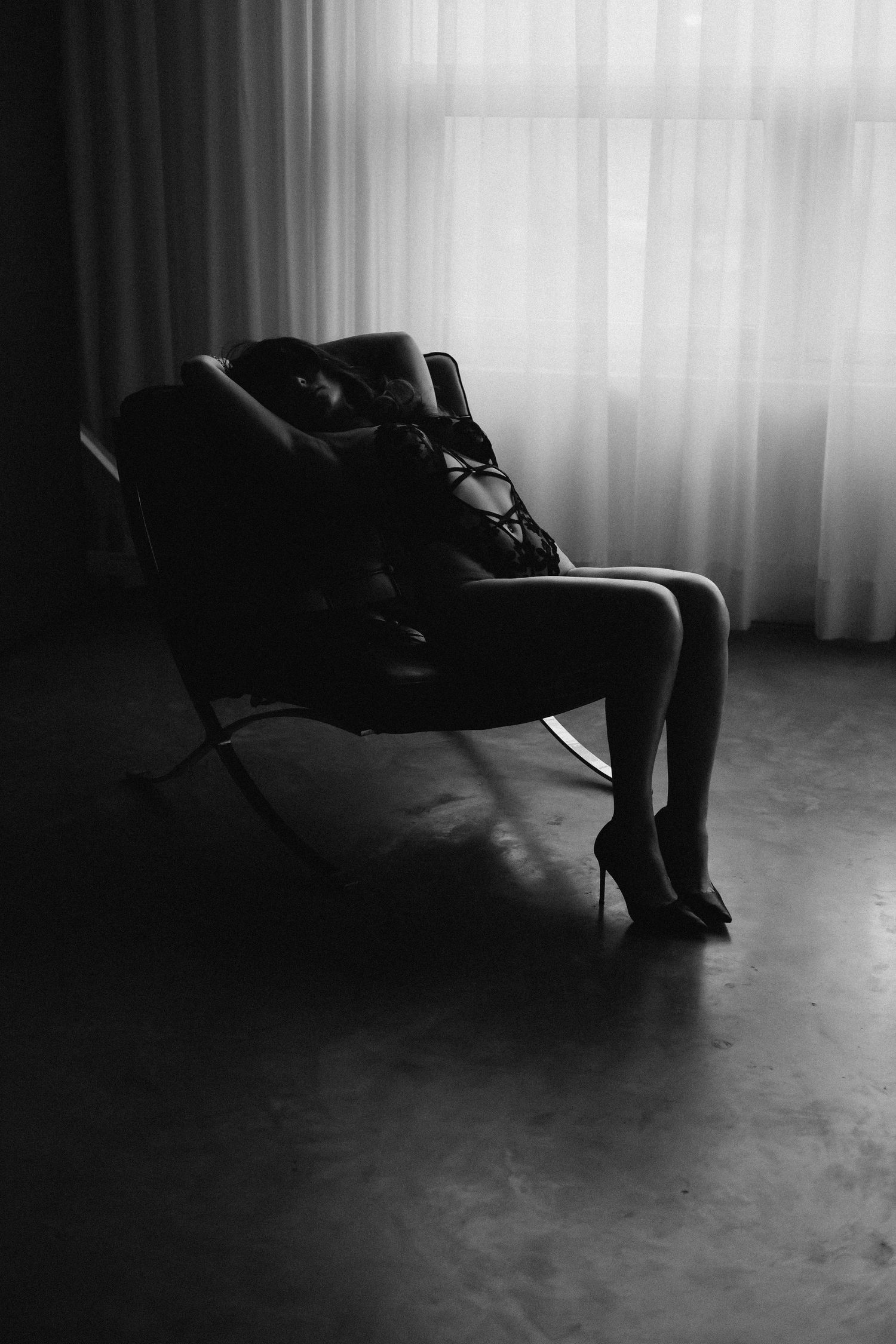 Black and white boudoir photo of a woman in heels reclining on a modern chair by sheer curtains.