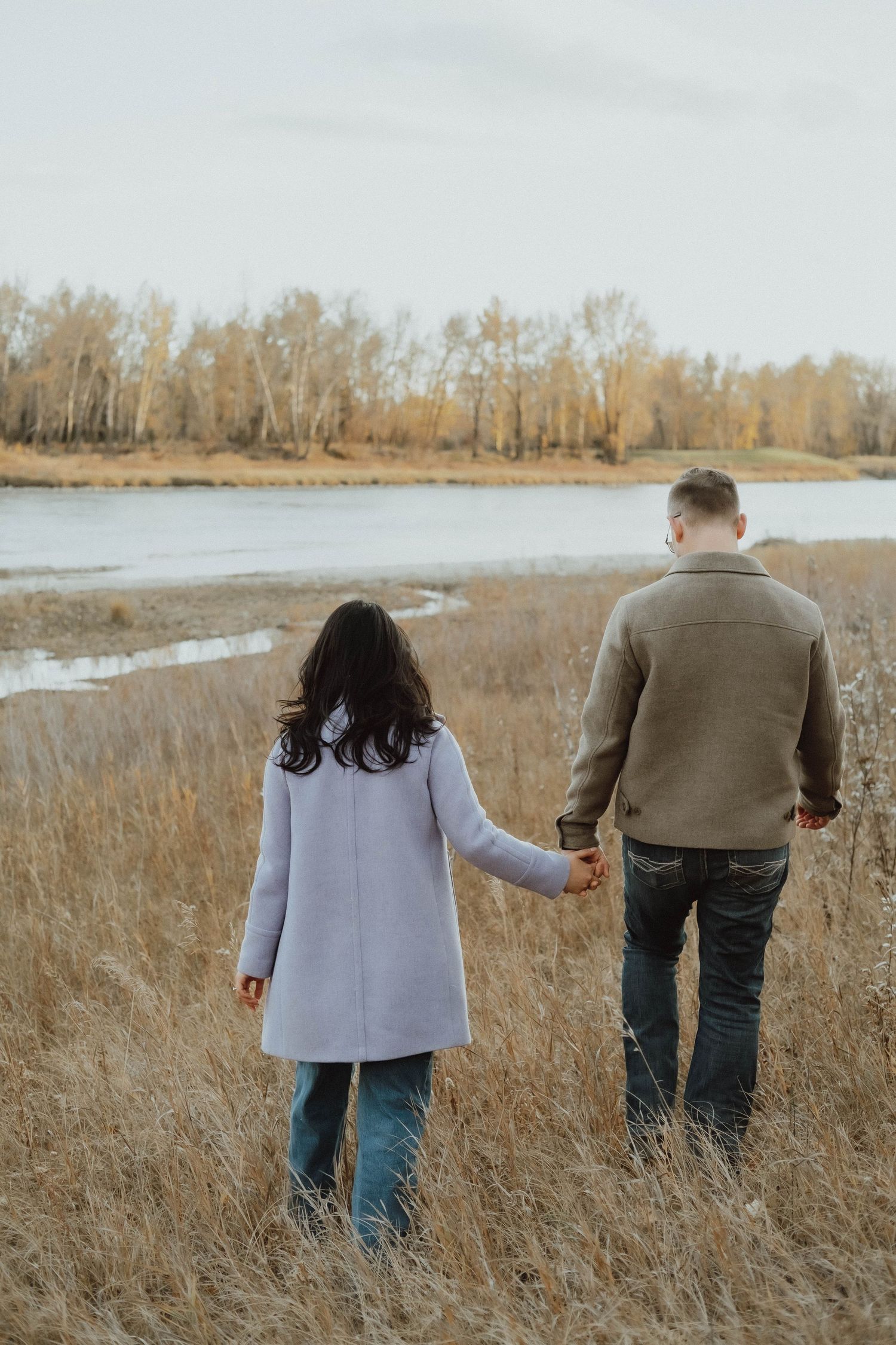 Couple holding hands walking through golden autumn field near a river with bare trees in background.