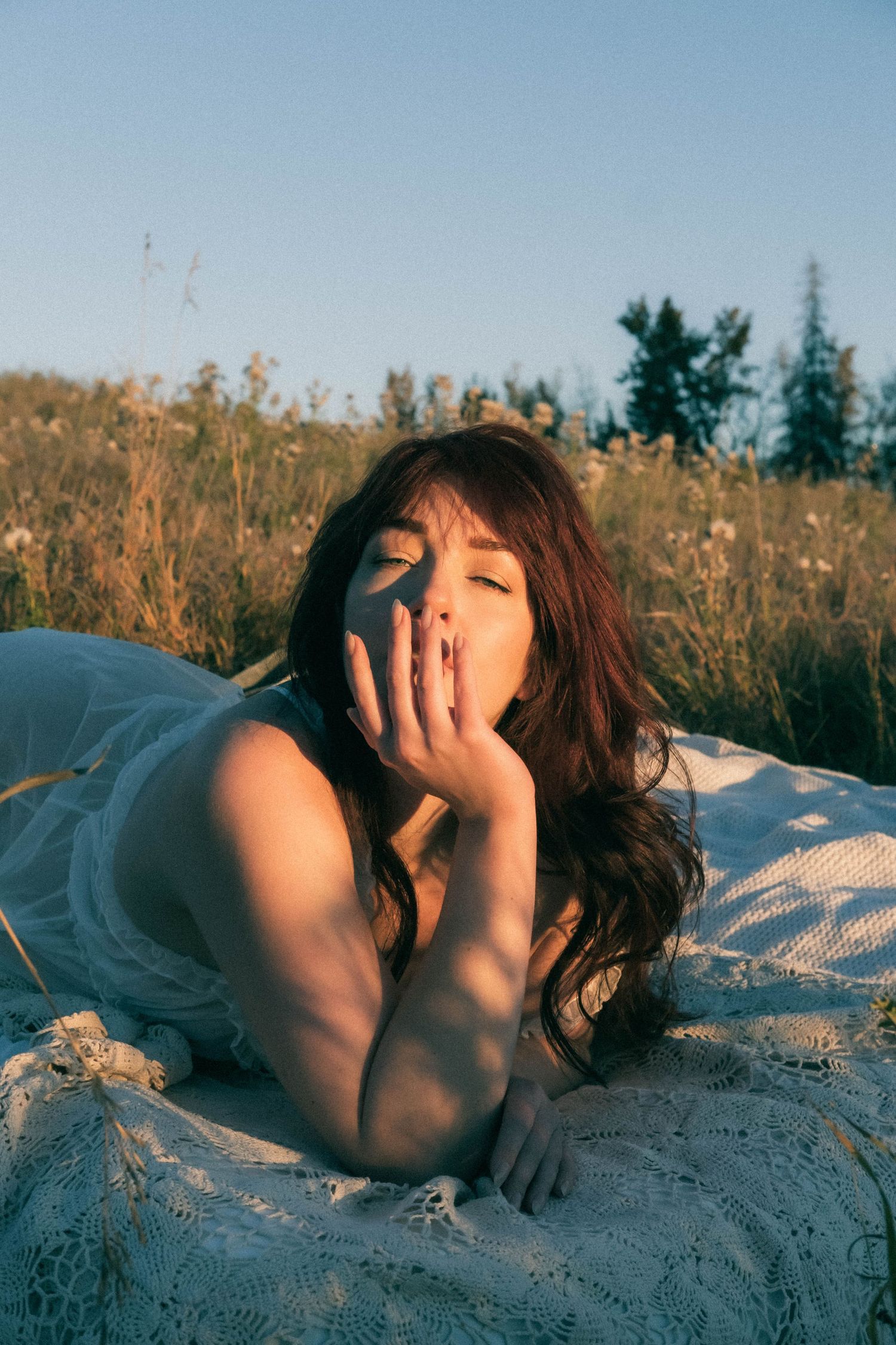 Woman with dark hair lying on white lace blanket in meadow, eyes closed, hand near face in golden sunlight.