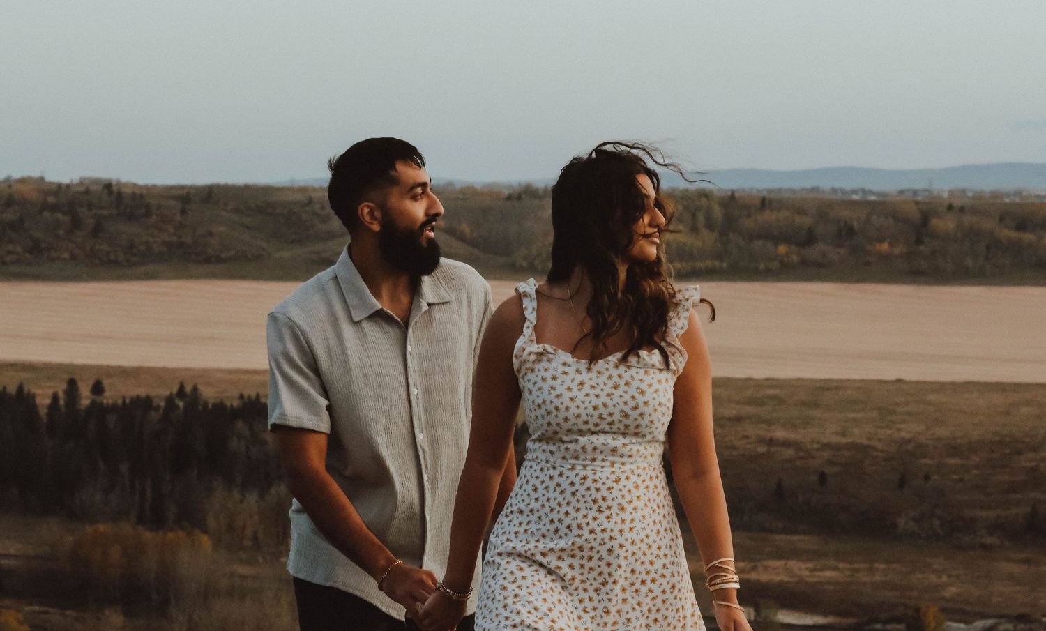 Couple holding hands outdoors at golden hour with scenic landscape and mountains in the background.