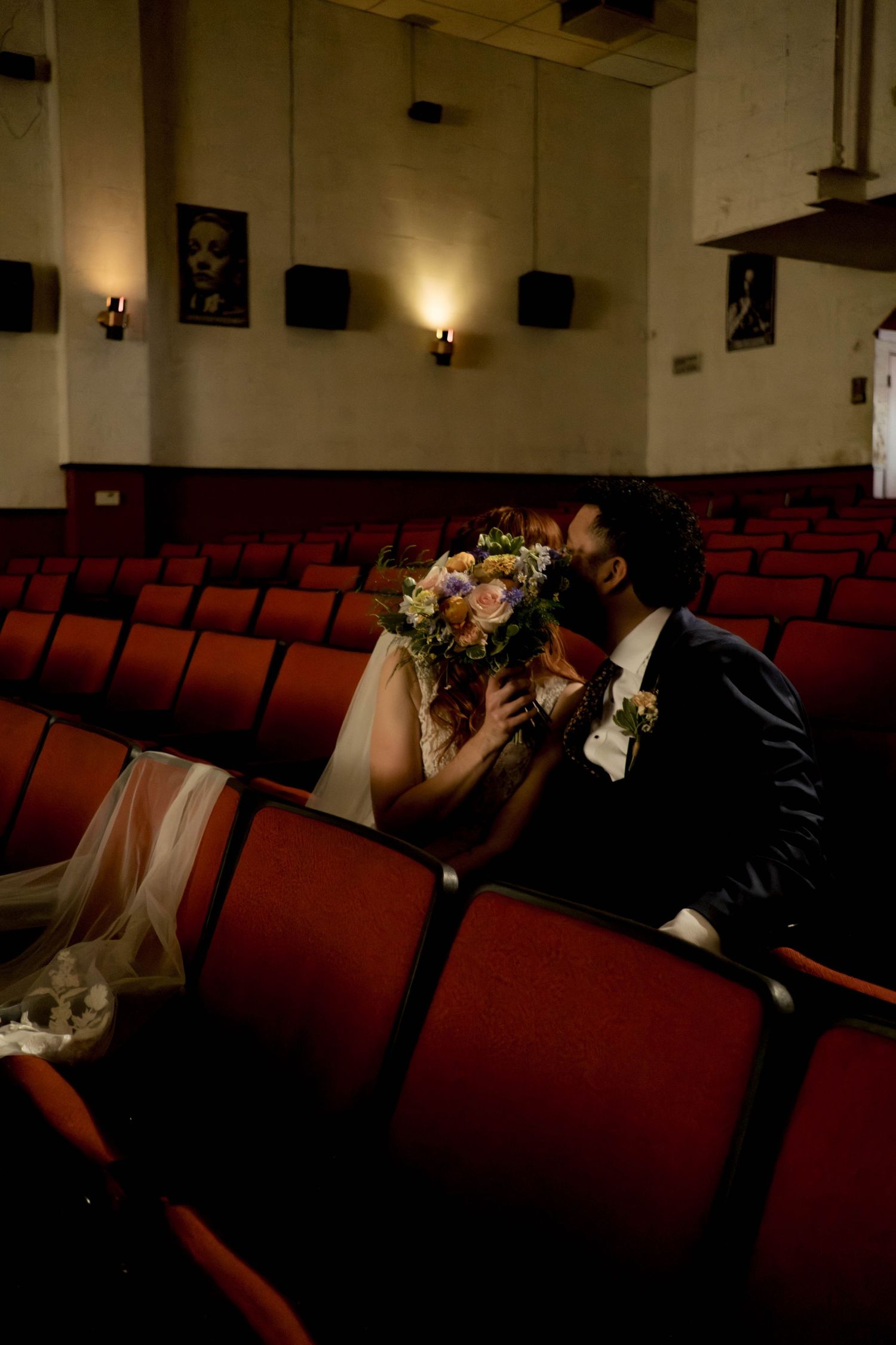 Bride and groom kissing behind a bridal bouquet while seated in red theater seats during their wedding.