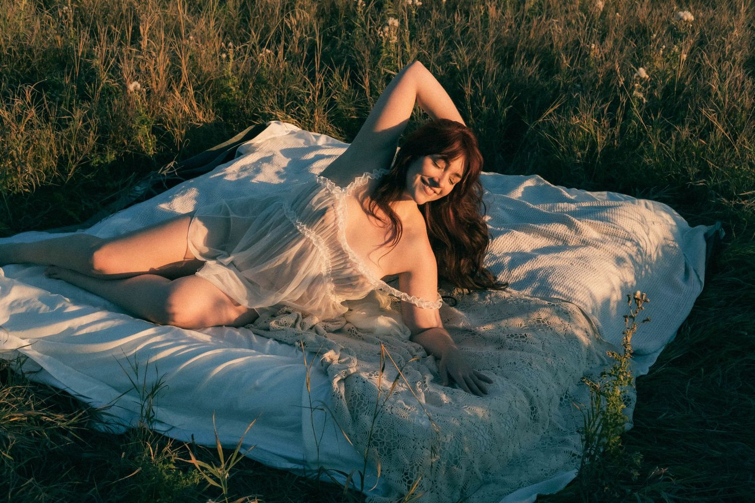 Woman in sheer white dress lying on mattress outdoors in golden hour sunlight surrounded by wild grass.