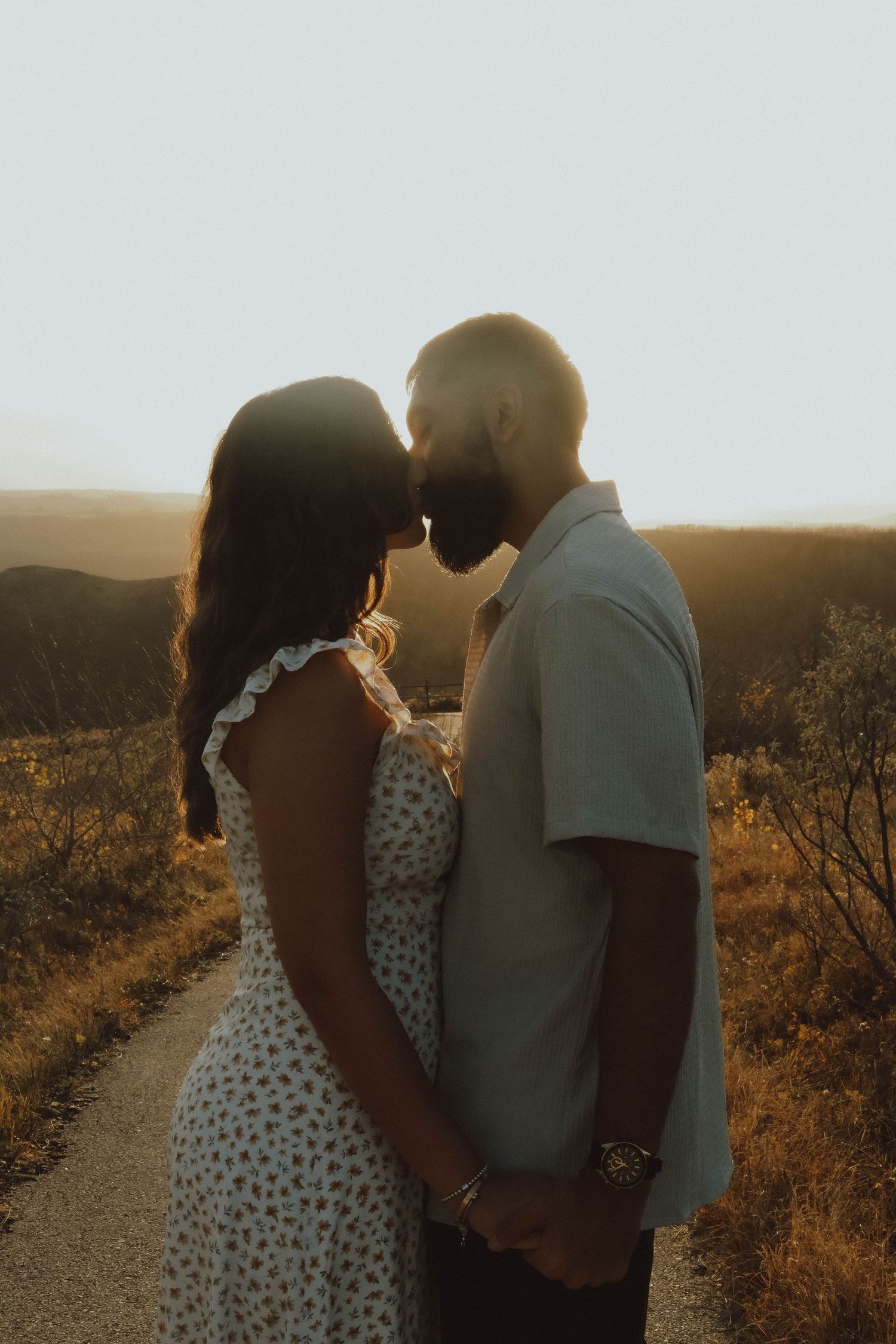 Couple sharing a romantic kiss at golden hour sunset on a scenic overlook path, woman in floral dress holding hands.