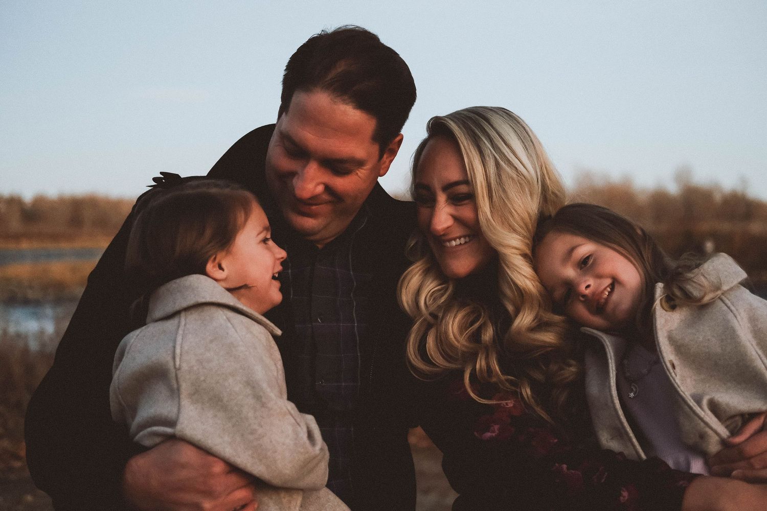 Happy family of four sharing a joyful moment outdoors near a lake during golden hour sunset.