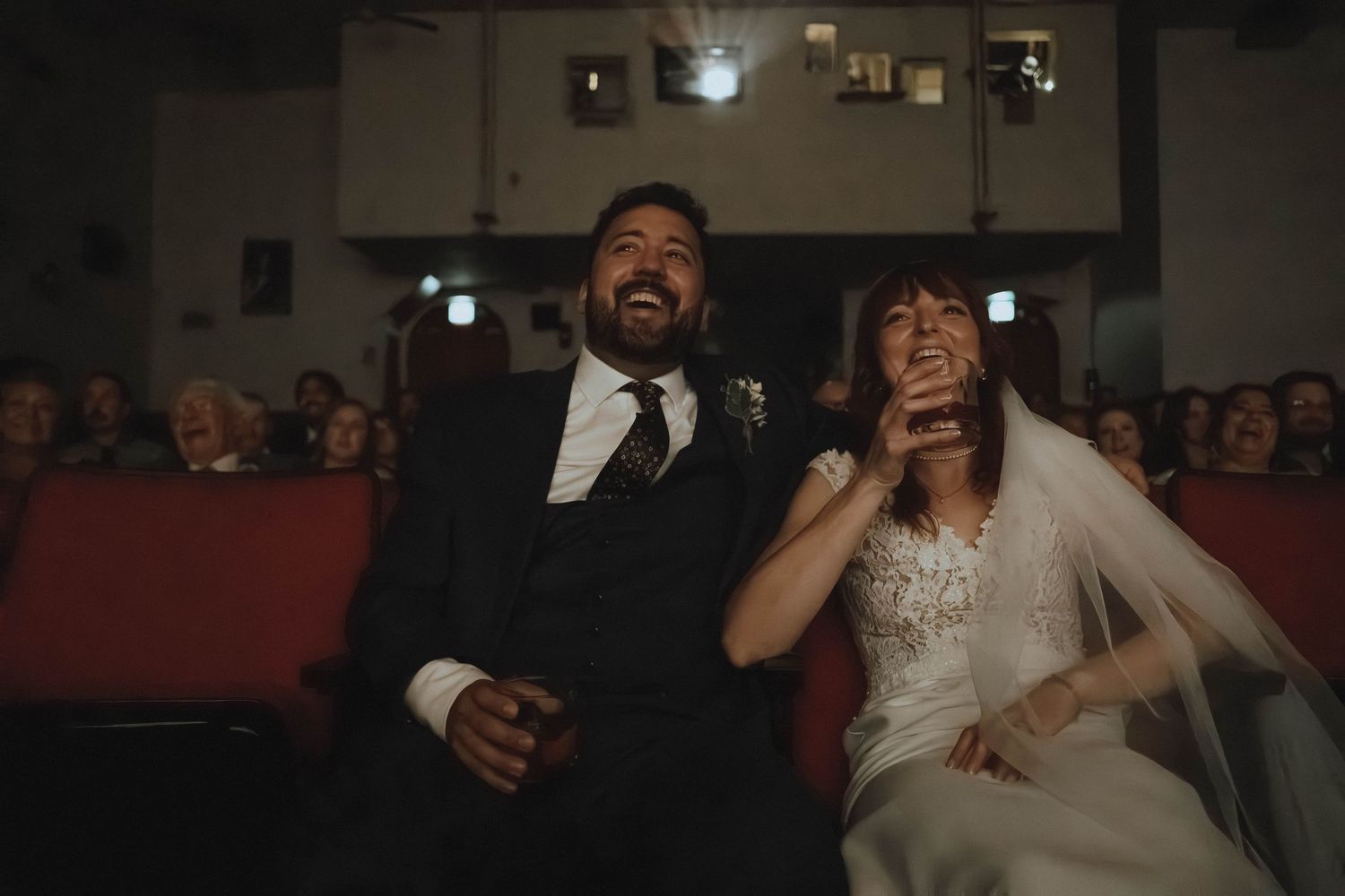 A joyful bride and groom laugh together during their wedding ceremony seated in a vintage theater venue.