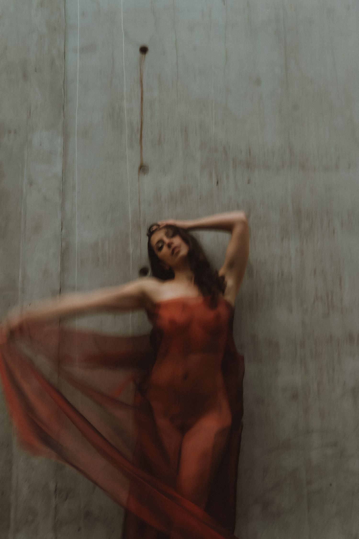 Dancer draped in sheer red fabric poses against concrete wall in artistic blur.