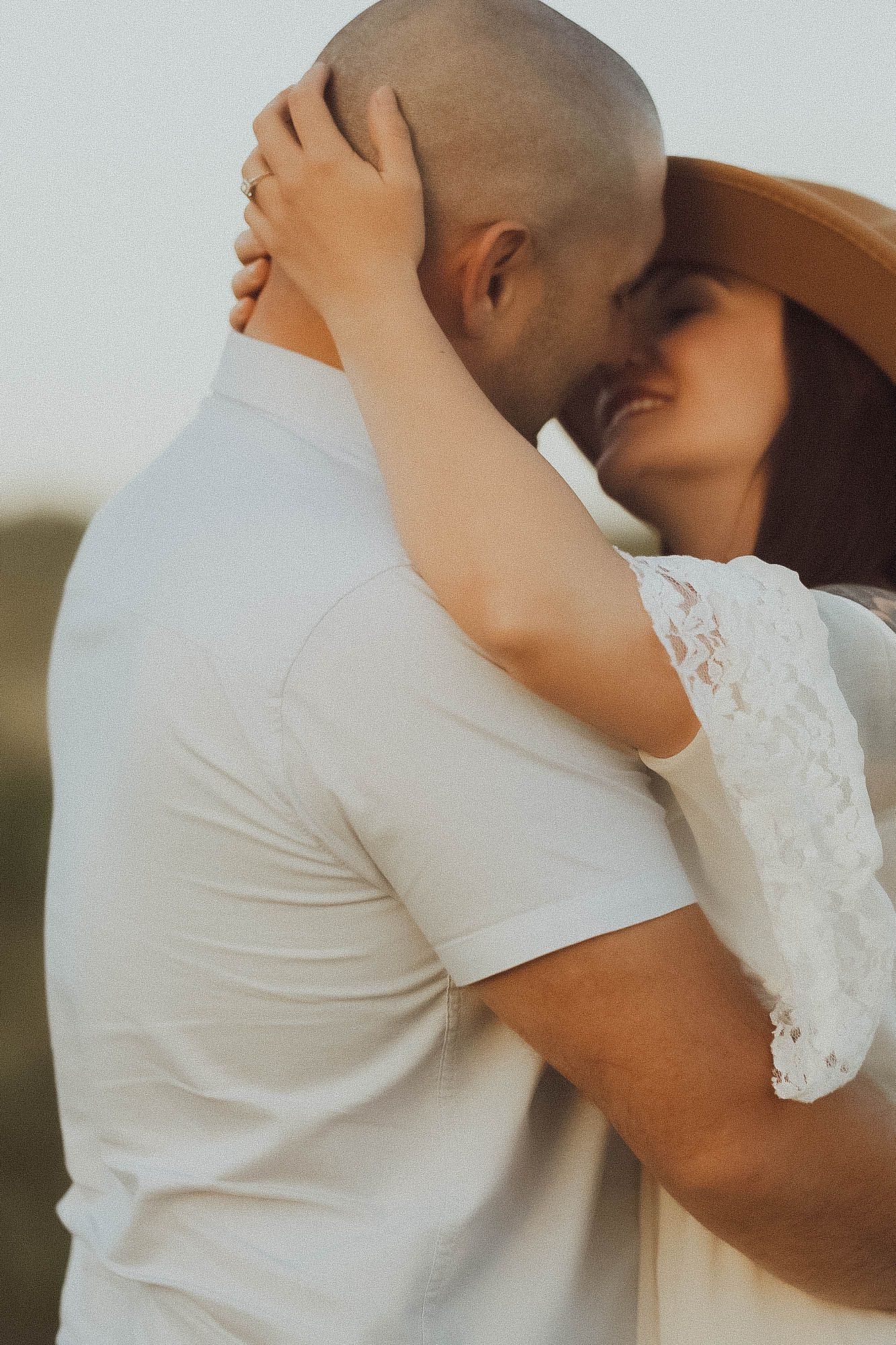 A couple sharing a tender embrace outdoors, woman in white lace dress and hat pulling man close for a kiss.